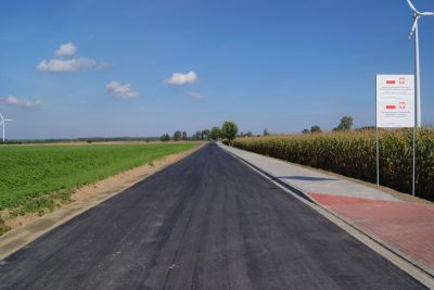 Droga asfaltowa, po lewej stronie pas zieleni, po prawej stronie chodnik, pola kukurudzy i tablica informacyjna, w oddali...
