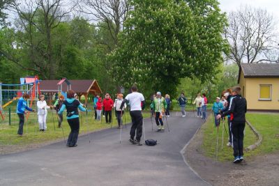 Grupa osób prowadzi rozgrzewkę z kijkami do nordic walking, w tle park i plac zabaw