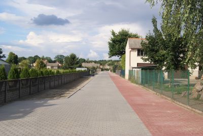 Droga z szarej kostki, z prawej strony pas drogi z czerwonej kostki. Po lewej stronie betonowy płot, za nim drzewa, w...