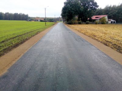 Droga asfaltowa, po obu stronach drogi pobocza gruntowe, po lewej stronie pas zieleni, po prawej stronie ścięte zboże, w...