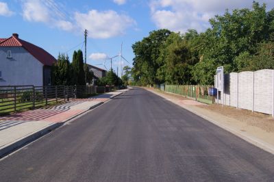 Droga asfaltowa, po lewej stronie chodnik i zabudowania, po prawej stronie płoty i drzewa