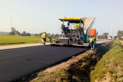 Prowadzone roboty drogowe, pas drogi asfaltowej, maszyna budowlana, robotnicy, po prawej stronie rów, w oddali łąka