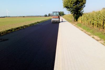 Chodnik z kostki brukowej i pas drogi asfaltowej, maszyna budowlana, po lewej stronie łąka, po prawej stronie drzewo i pole...