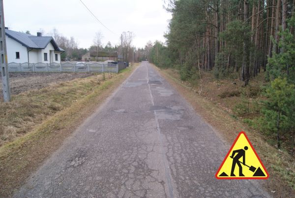 Pas drogi o nawierzchni asfaltowej, z lewej strony biały budynek ogrodzony betonowym płotem i pola, z prawej strony las. W...