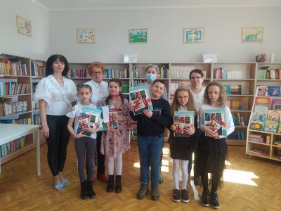 Na zdjęciu grupa osób: cztery panie z jury i pięcioro dzieci z dyplomami i nagrodami książkowymi. W tle regały z książkami.