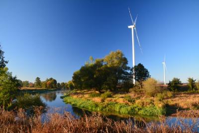 Zakole rzeki w słonecznej, jesiennej scenerii. W tle dwa wiatraki do produkcji energii elektrycznej.